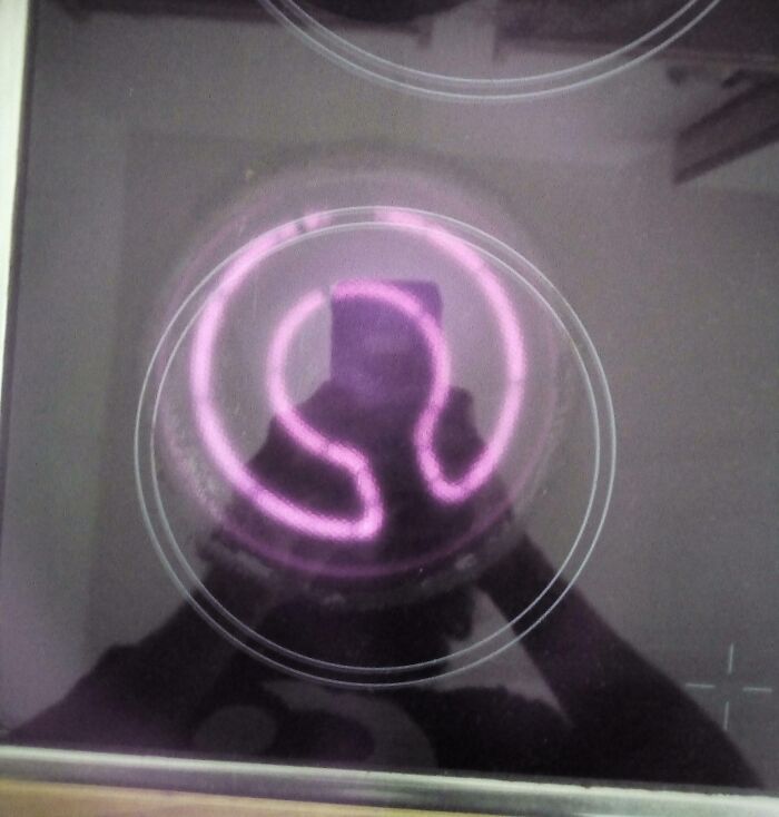 Reflection of a person taking a photo of a glowing purple electric stove burner on a smooth cooktop surface, imperfect details visible.