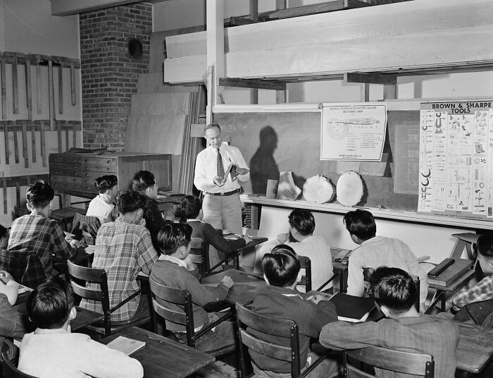 Teacher instructing students in a woodworking classroom in a school during World War II with tool charts on the wall.