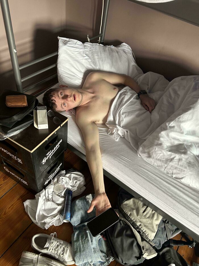 Young man reaching for his phone from a messy floor, lying in bed, captured in accidental renaissance photo style.