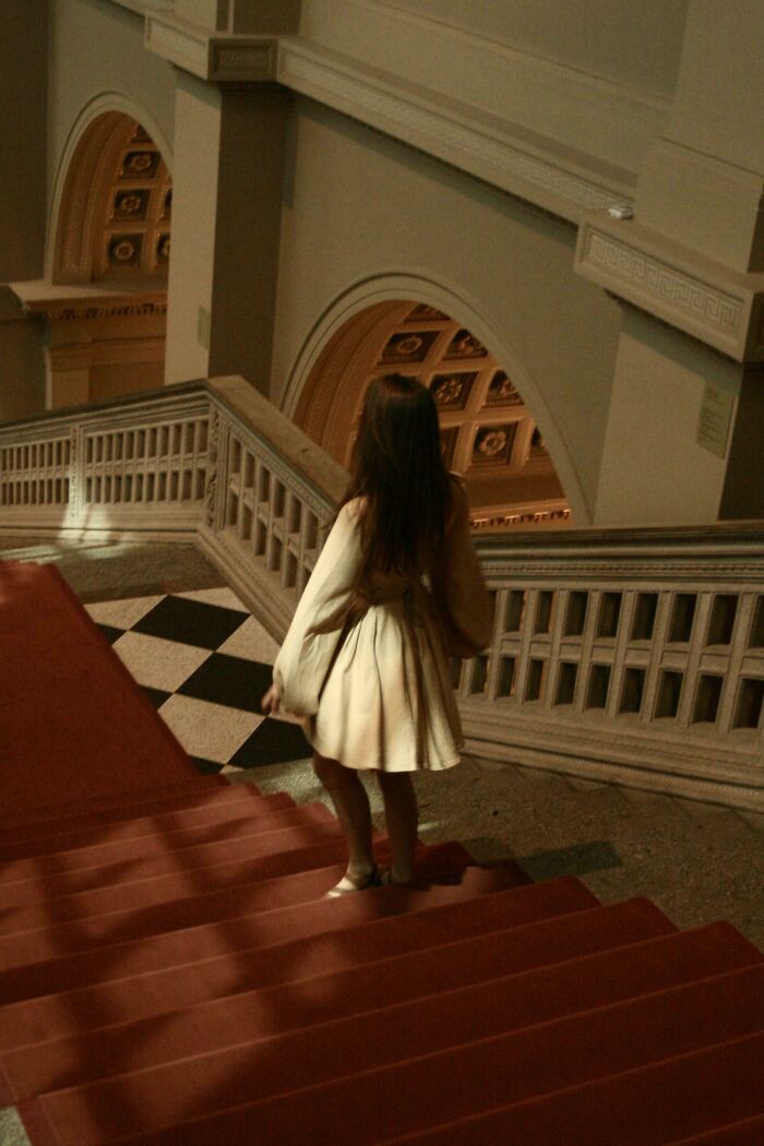 Woman in a flowing dress descending grand staircase in historic building resembling a Renaissance masterpiece scene.