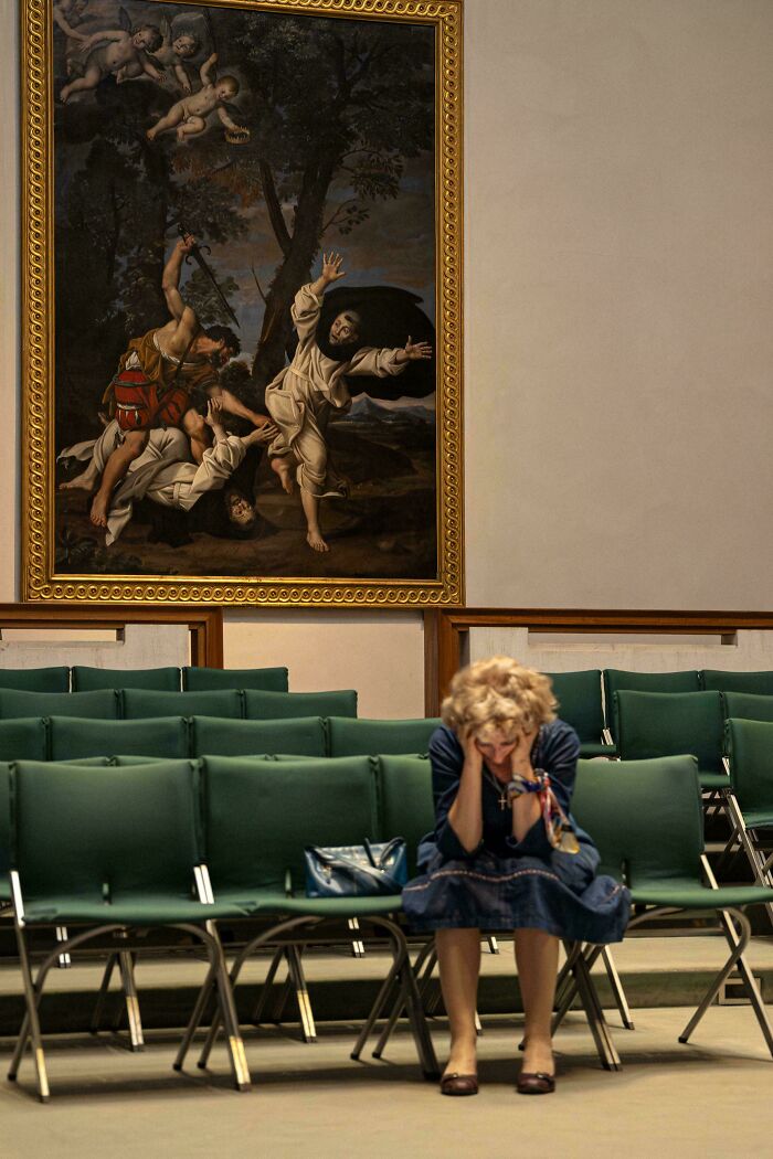 Woman sitting with head in hands below large accidental renaissance photo displayed in a museum setting with green chairs.