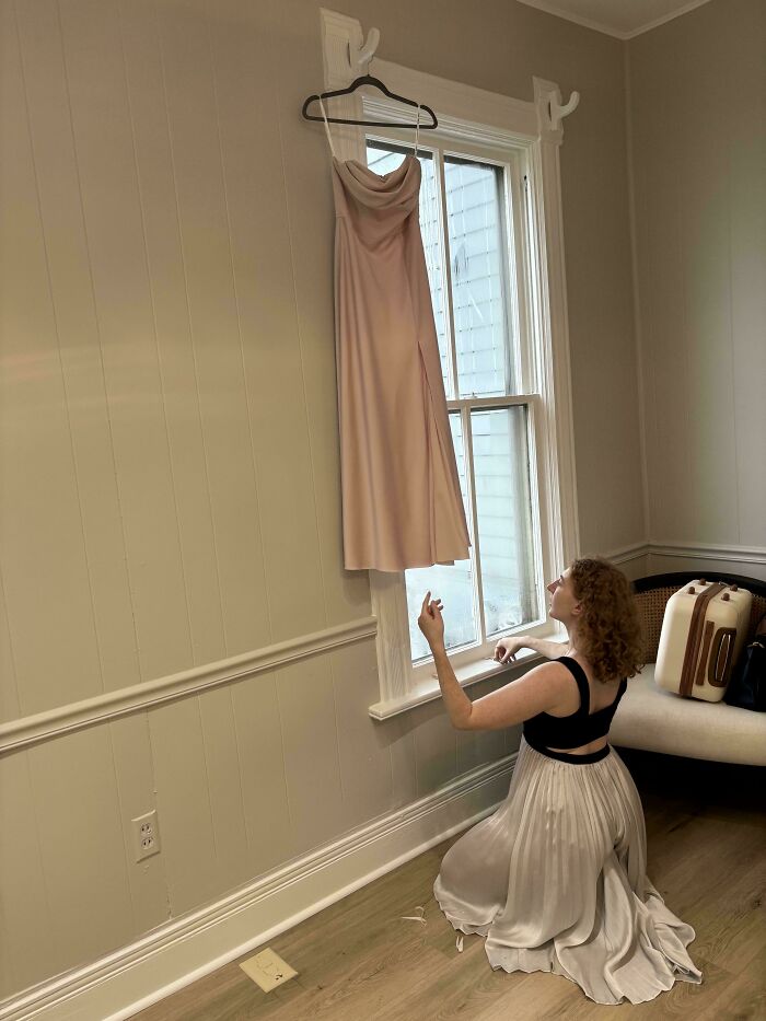Woman in light dress kneeling by window, reaching towards hanging gown, an accidental Renaissance photo moment.