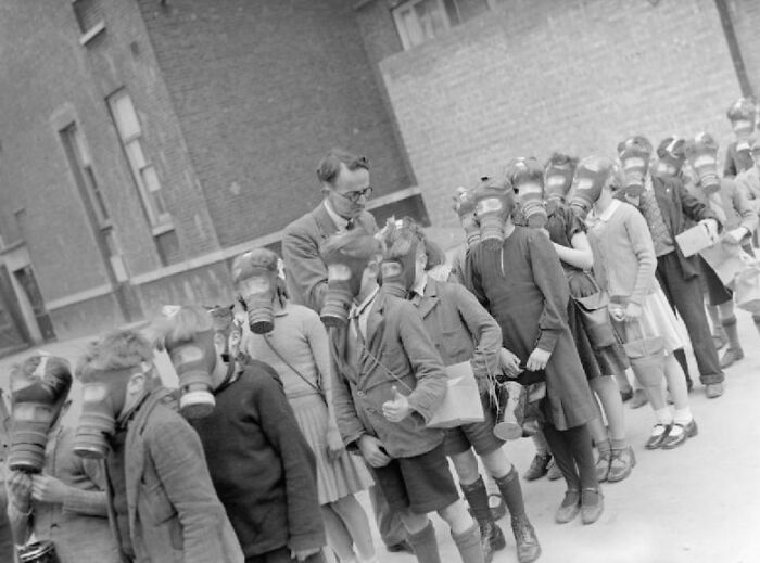 Schoolchildren wearing gas masks lined up outdoors for a World War II safety drill at a school during wartime.