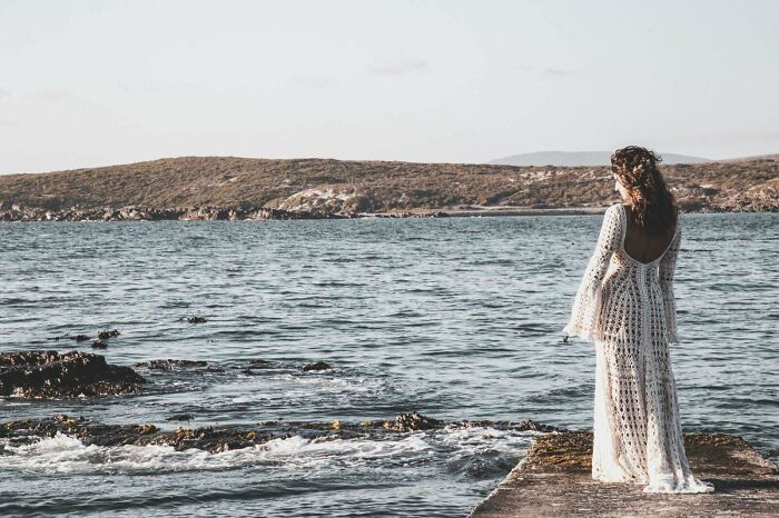Woman wearing a crocheted dress standing on a pier overlooking the sea with hills in the background, showcasing crochet craftsmanship.