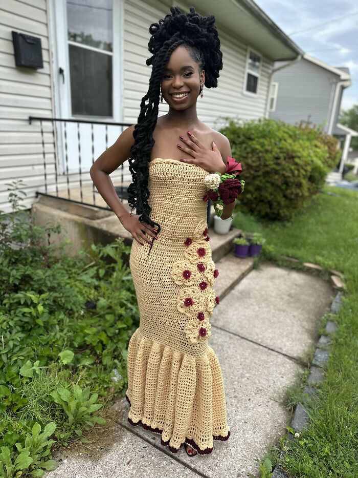 Young woman wearing an intricate handmade crochet dress adorned with floral details, smiling outdoors in a garden setting.