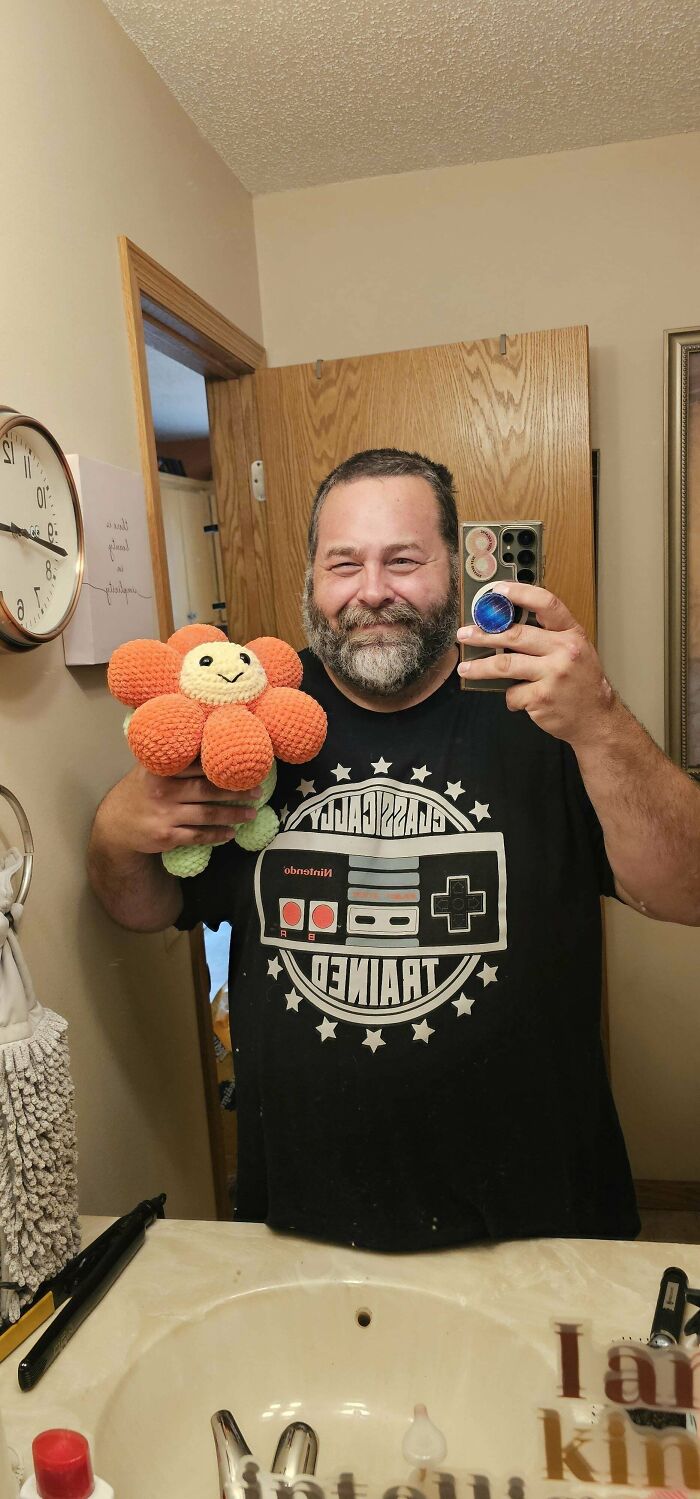 Man smiling in bathroom mirror holding a crocheted orange flower, showcasing incredible crochet craftsmanship and creativity.