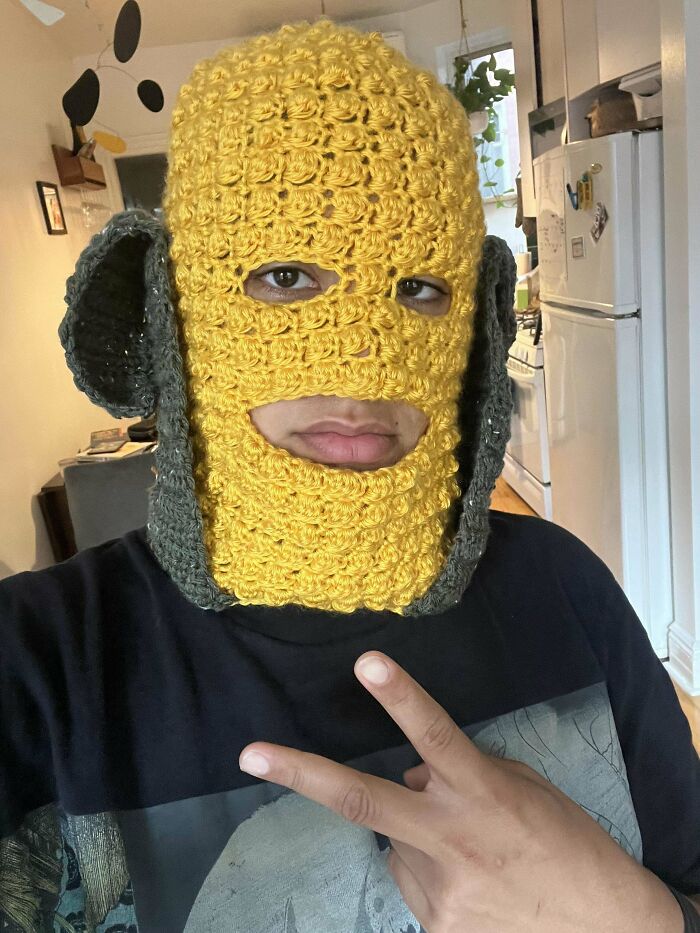 Person wearing a yellow and gray crocheted mask with eye and mouth holes, showcasing incredible crochet craftsmanship indoors.