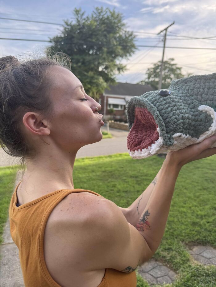Woman holding and kissing an intricate crochet fish puppet outdoors, showcasing impressive crochet craftsmanship.