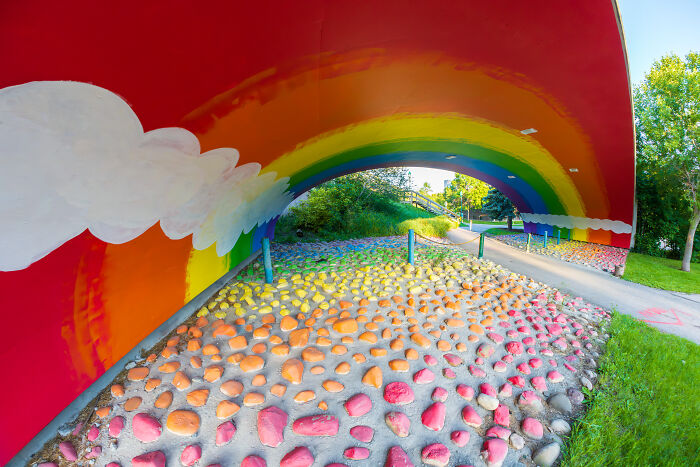 Colorful rainbow mural under a bridge with painted stones on the ground, capturing a modern life boring dystopia scene.