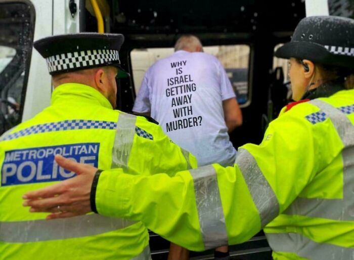 Two Metropolitan Police officers escort a man wearing a T-shirt with a political message in a boring dystopia scene.