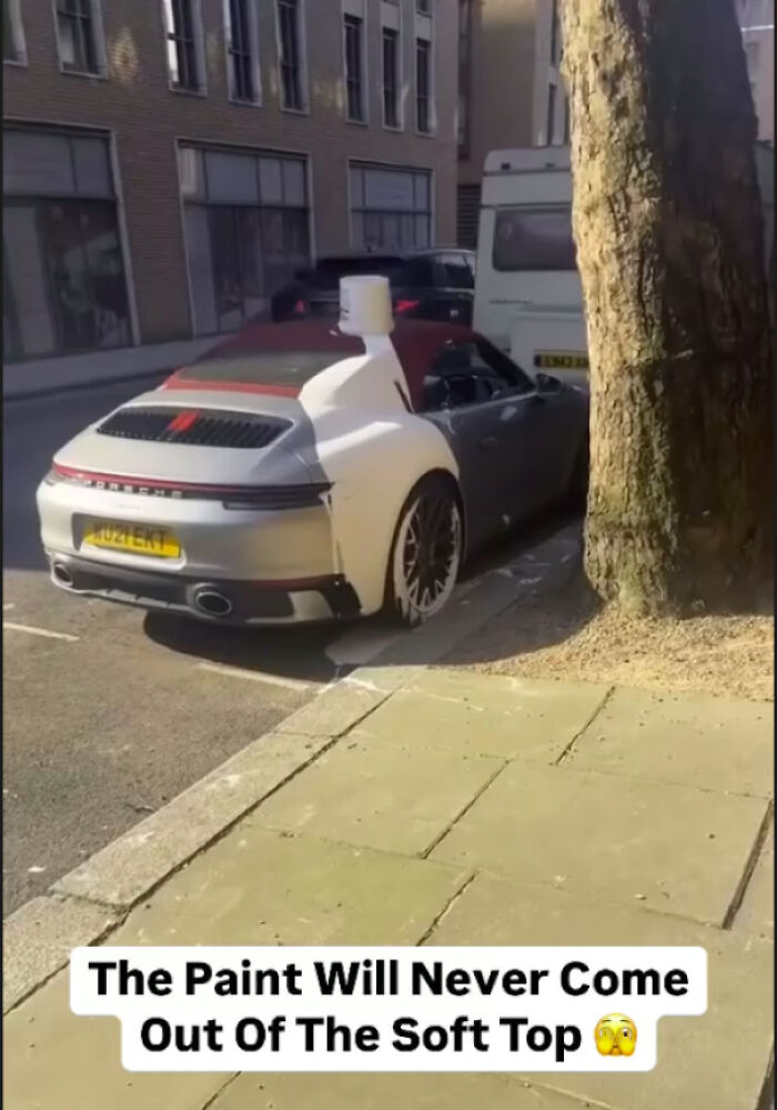 A white Porsche with an open red soft top and white paint splatters parked next to a tree on the street. A true accident that looks expensive.