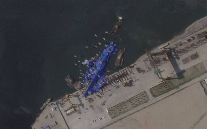 Aerial view of a blue-covered shipwreck in a harbor, an expensive accident, with cranes and barges nearby.