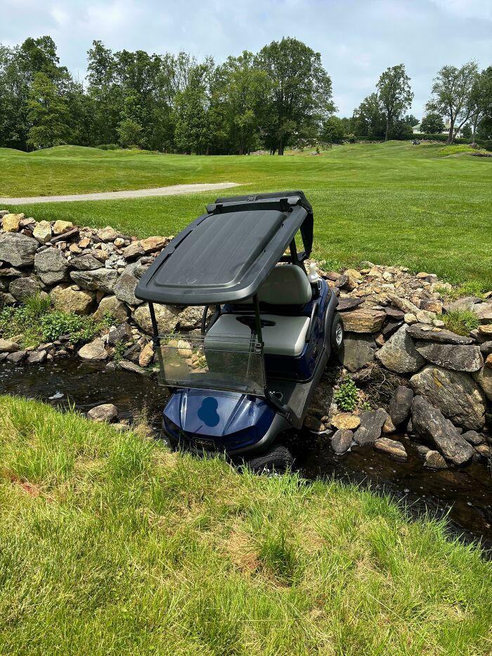 A blue golf cart in a stream next to a stone wall on a sunny golf course, an accident. Fails that looked expensive.