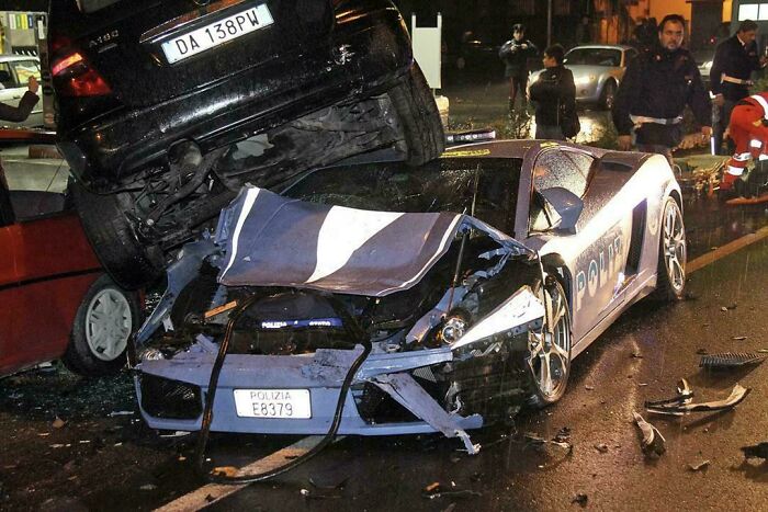 A shocking fail: a black car rests atop a wrecked police Lamborghini, surrounded by debris. Bystanders and emergency services are present, inspecting the scene.