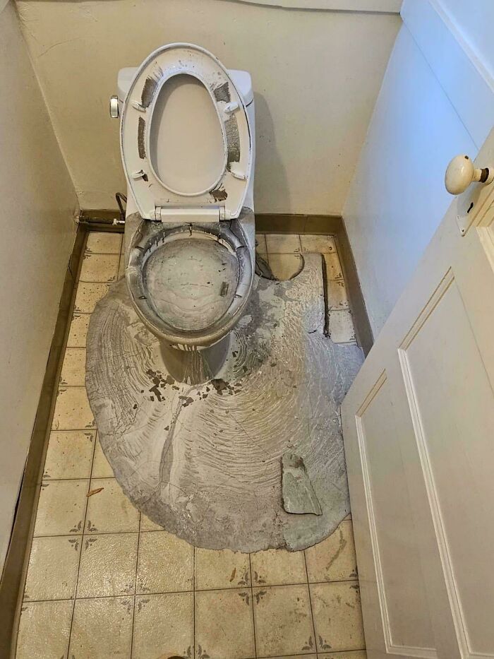A damaged toilet sits in a small bathroom, surrounded by a large spill of cement-like debris. A real accident.