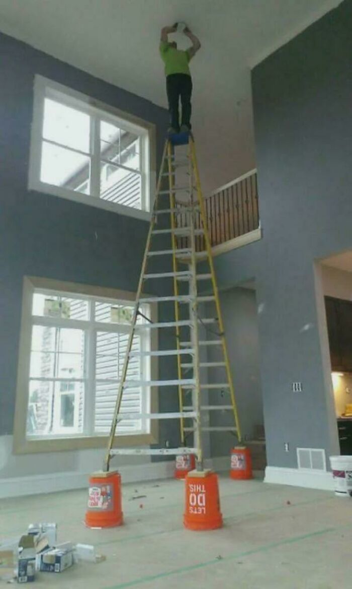 Employee blatantly ignoring safety protocol standing on unstable ladder setup with buckets in a high-ceiling residential space.