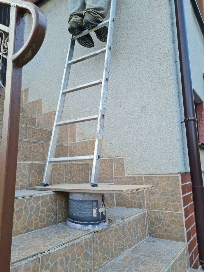 Worker ignoring safety protocol by standing on unstable ladder setup on stairs with plank and bucket support