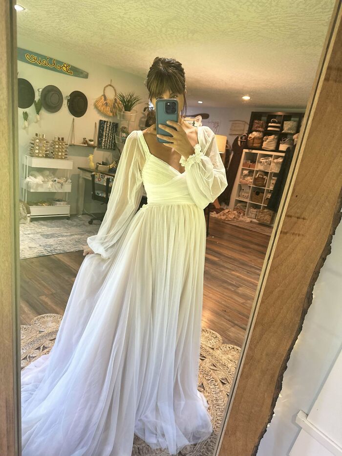 Woman wearing a flowing white dress with sheer sleeves taking a mirror selfie in a cozy room showcasing cool creations.