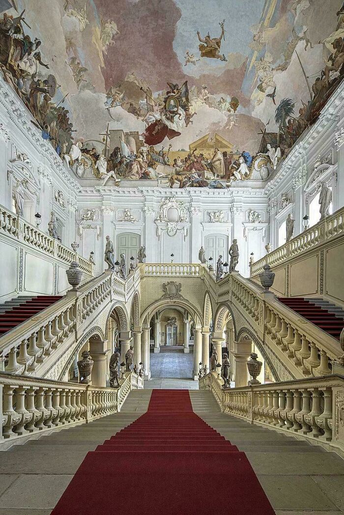 Grand staircase with red carpet and detailed sculptures in a historic building showcasing impressive architectural feats.