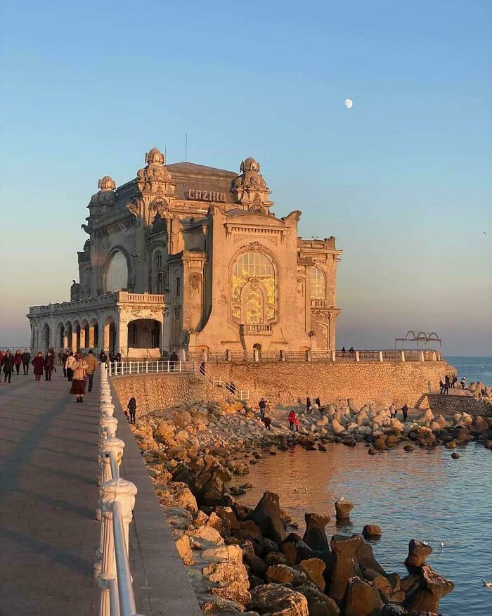 Historic architectural feats by the sea at sunset with people walking on a pier beside rocky shoreline and calm water.