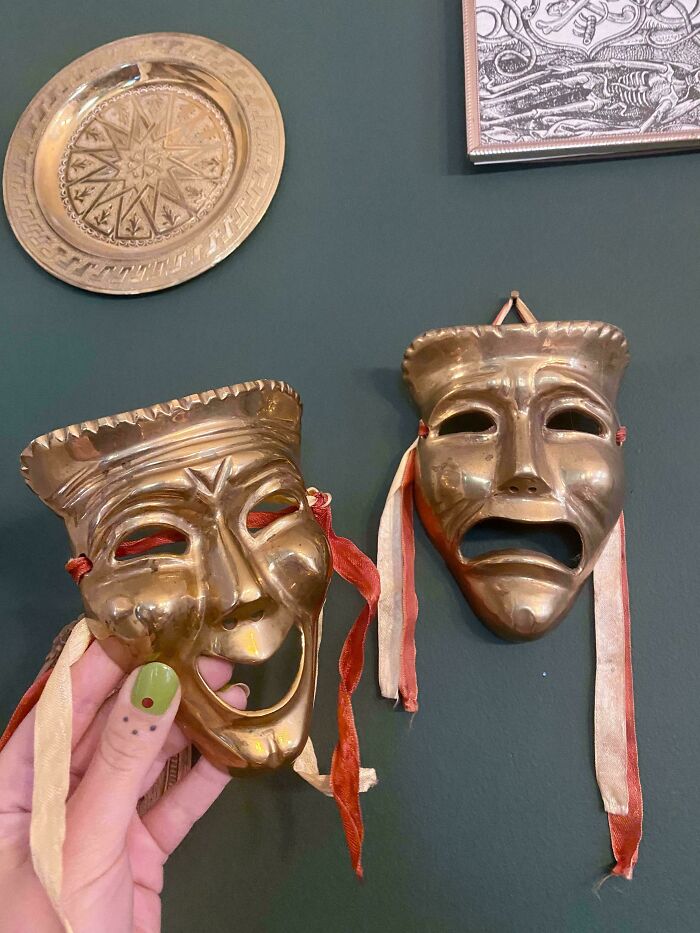 Two vintage brass theatrical masks with ribbons, one held by a hand, displayed as thrift store finds on a dark wall.