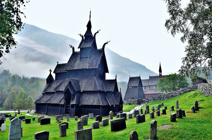 Dark wooden stave church surrounded by gravestones, set against misty mountains, showcasing eerie evil buildings architecture.
