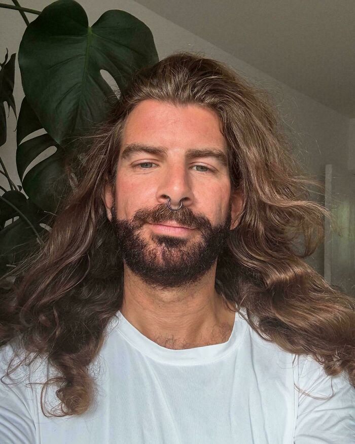 Man with fierceflow long beautiful hair and beard, wearing a white shirt, posing indoors with green plant in background.