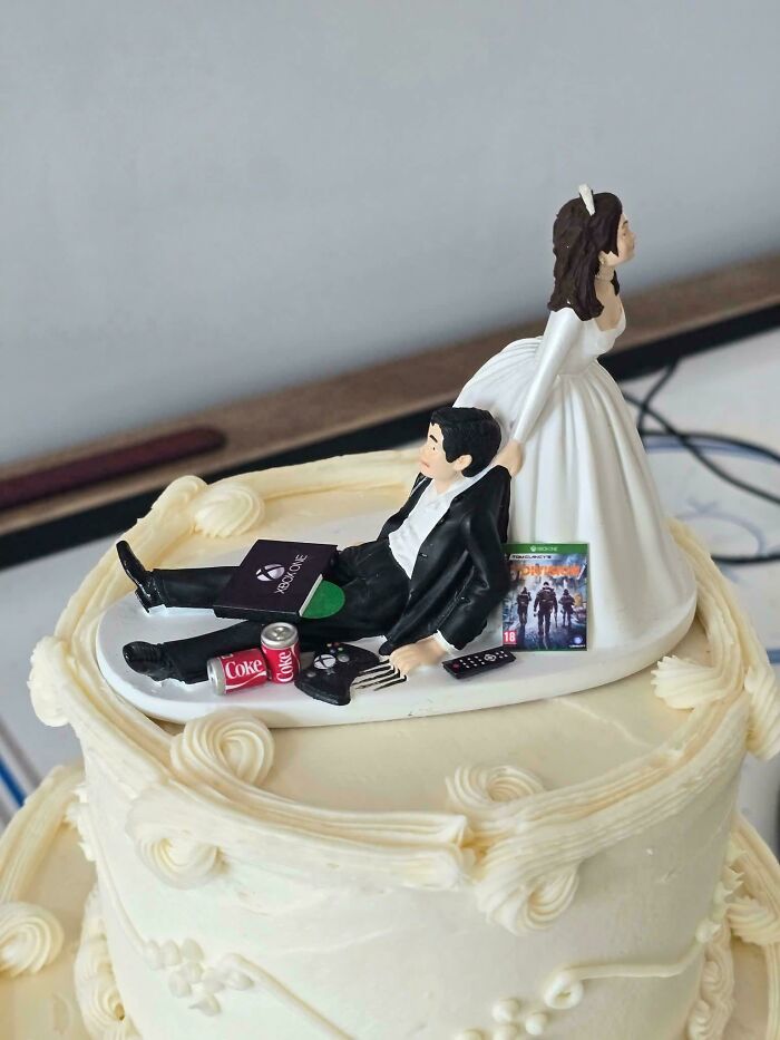 Wedding cake topper showing a bride pulling a groom distracted by video games and soda, highlighting worst wedding guests.