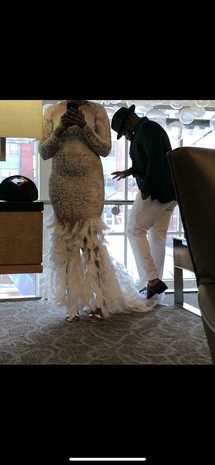 Person in an embellished wedding dress with feathers standing next to a man in white pants and black jacket indoors.