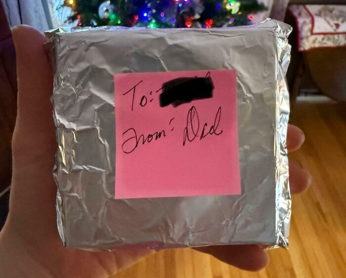 Hand holding a small gift wrapped in foil with a pink note labeled from Dad, showcasing hilarious dad creativity.