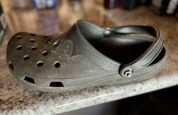 Worn black croc shoe with holes and a bolt attached, showcasing a hilarious dad's inventive fix for fatherhood.