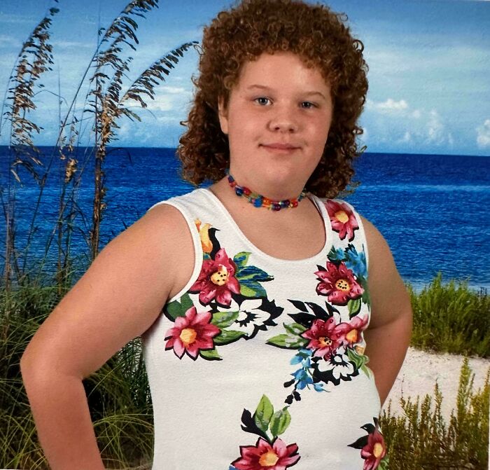 Child with curly hair wearing a floral tank top and beaded necklace posing in front of a beach backdrop in a regrettable past pic.