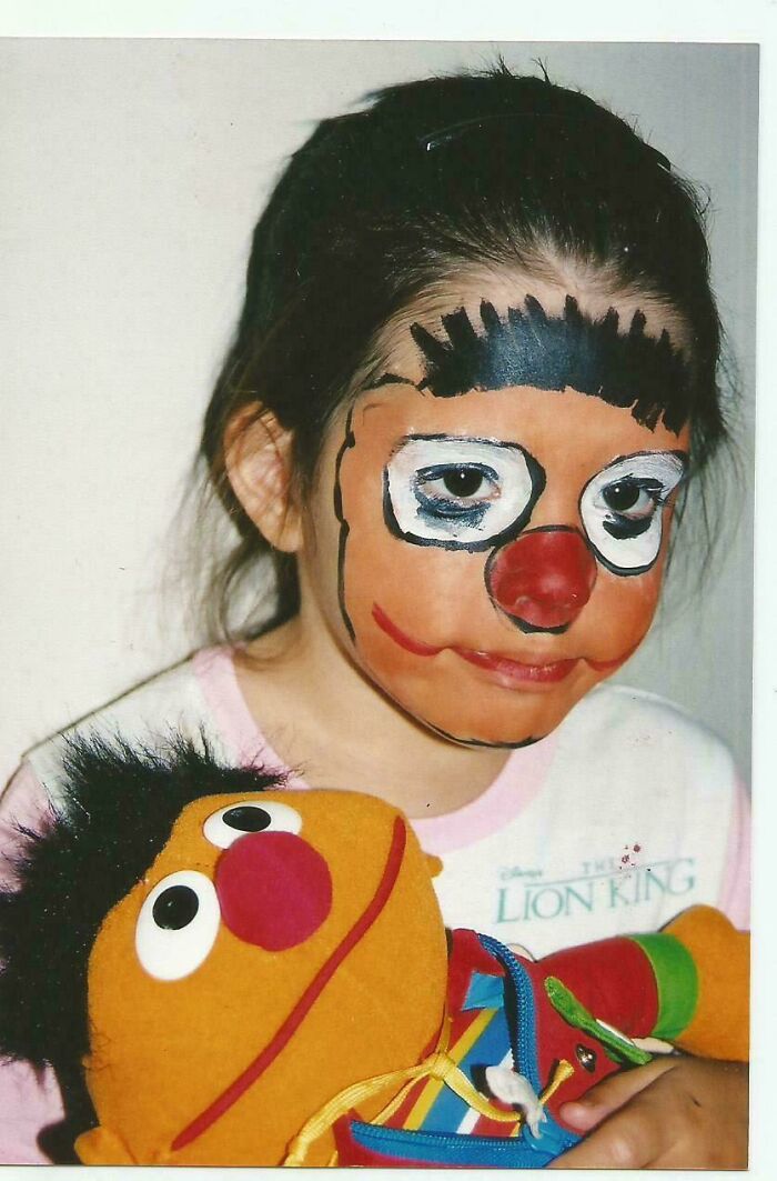 Child with clown face paint holding a colorful Ernie doll, a cringe-worthy moment from people’s regrettable past photos.