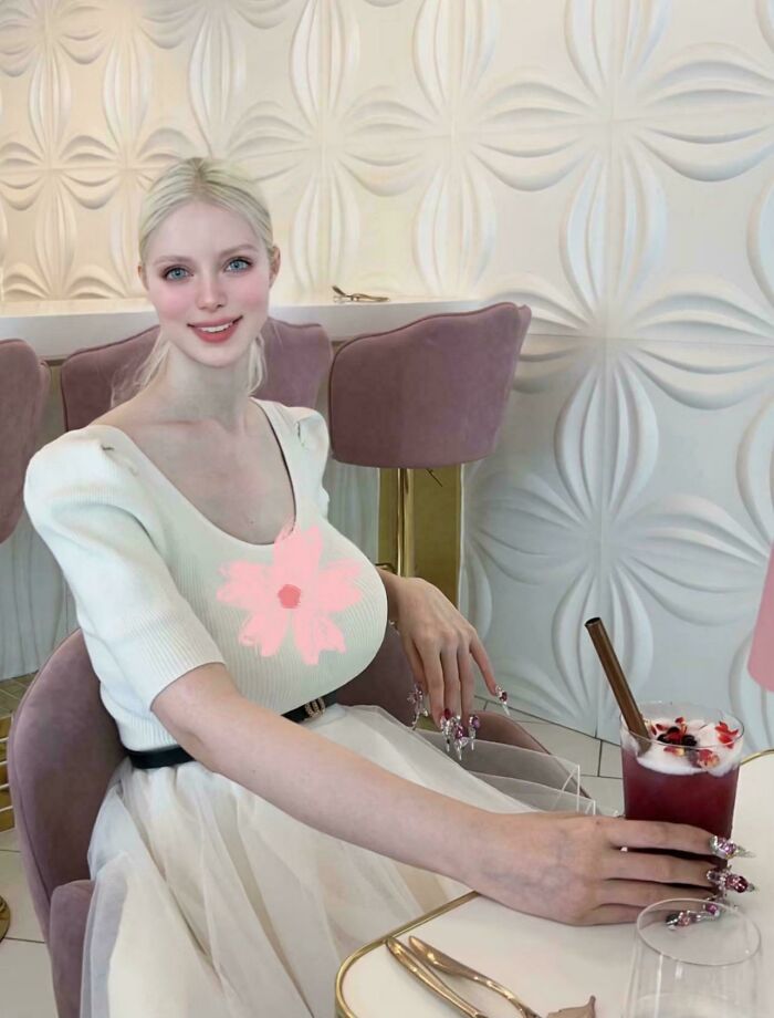 Young woman with exaggerated features sitting in a cafe holding a drink, illustrating Instagram vs. reality pics contrast.