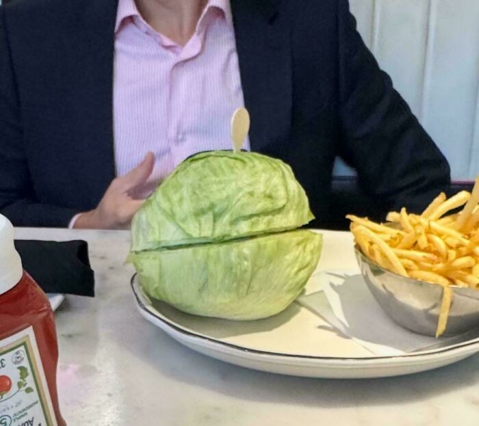 A person seated at a table with a burger wrapped entirely in lettuce and a bowl of French fries nearby.
