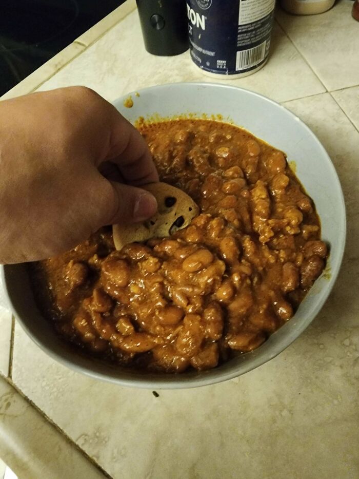 Hand dipping a chocolate chip cookie into a bowl of baked beans, an unusual dish combination.