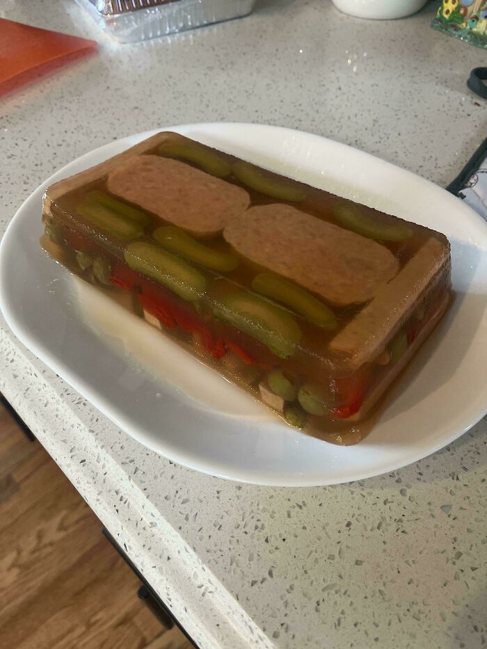 Unappetizing dish with sliced lunch meat and pickles set in gelatin on a white plate on a kitchen countertop.