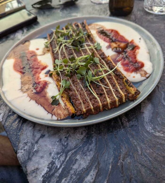 Unusual dishes with grilled textured food, drizzled sauce, garnished with microgreens on a grey plate outdoors.