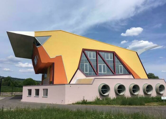 Bizarre building with unique geometric shapes and circular windows surrounded by grass under a blue sky with clouds.