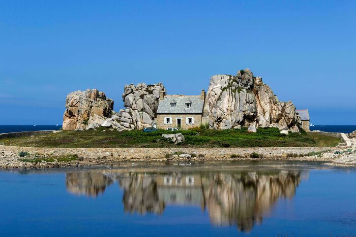 Unique bizarre building nestled between large rocks by calm water under a clear blue sky with natural reflection.