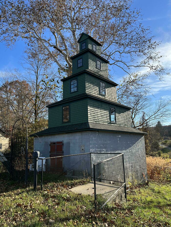 Bizarre building with stacked green shingled levels and a concrete base surrounded by a fenced yard under blue sky.