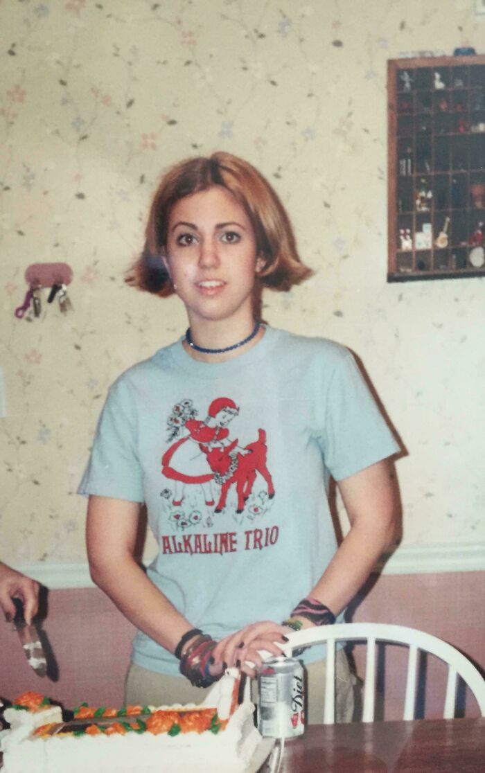 Teen girl wearing an Alkaline Trio shirt with bracelets and short hair, standing by a birthday cake and soda can indoors.