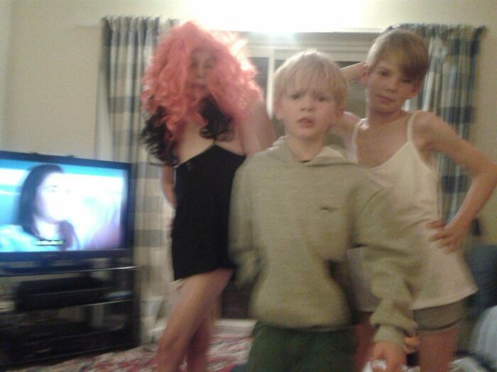Three kids in a living room, one wearing a pink wig and black dress, captured in a blurry regrettable past photo.