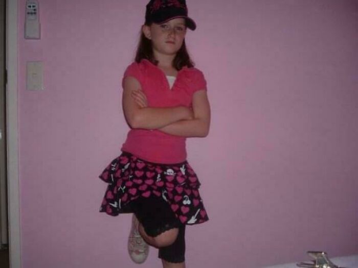 Young girl in a pink shirt and heart-patterned skirt posing confidently against a pink wall, a regrettable past photo cringe moment