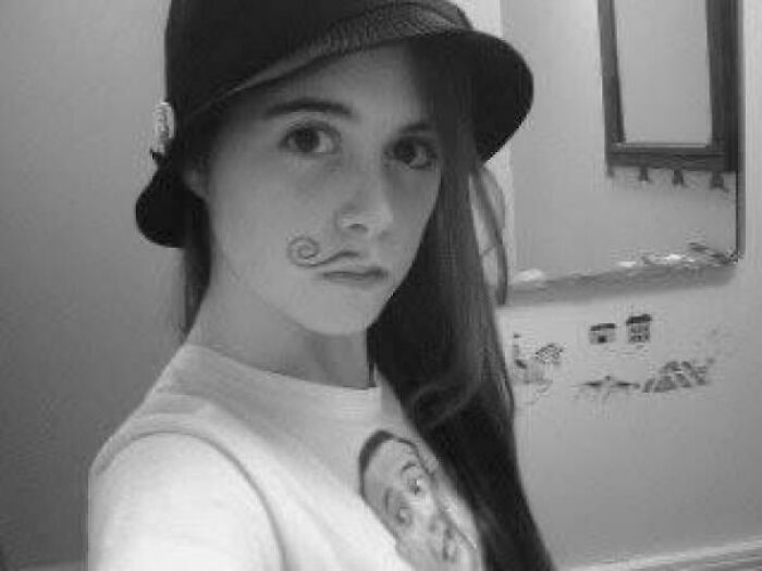 Black and white selfie of a young woman with a drawn-on curly mustache and a bucket hat, from someone's regrettable past pics.