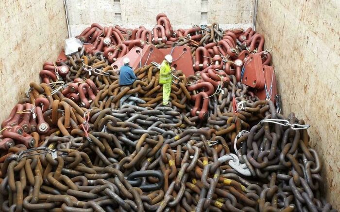 Two workers standing amidst a vast pile of extremely large chains, highlighting the scary and eerie scale.