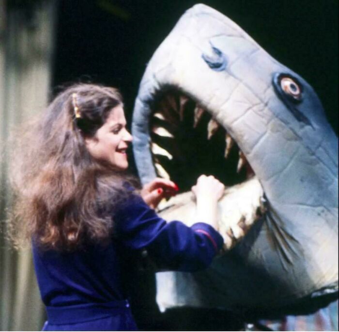 Woman in a blue outfit smiling and interacting with a large shark puppet in a nostalgic scene that makes people feel very old.