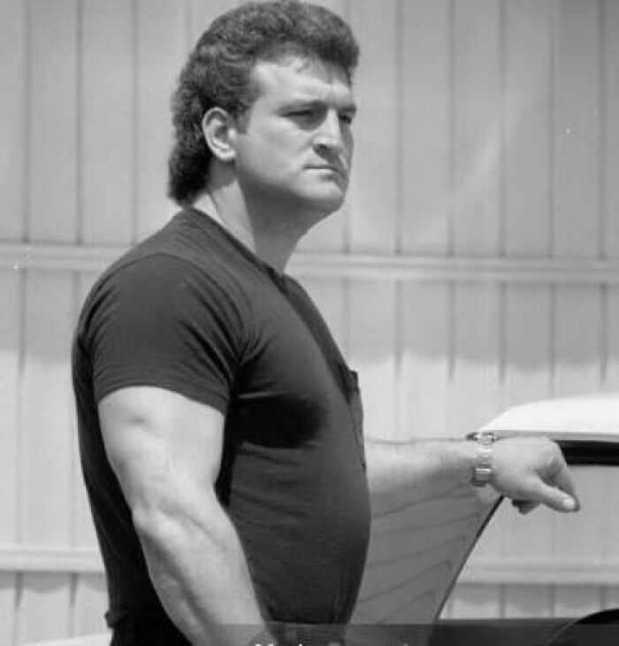 Man with 80s hairstyle and black shirt standing by a car, a nostalgic image for those who recognize and feel very old.