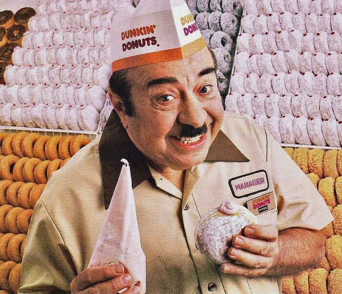 Dunkin Donuts worker holding a powdered donut and frosting bag surrounded by vintage donut displays, nostalgic food items.
