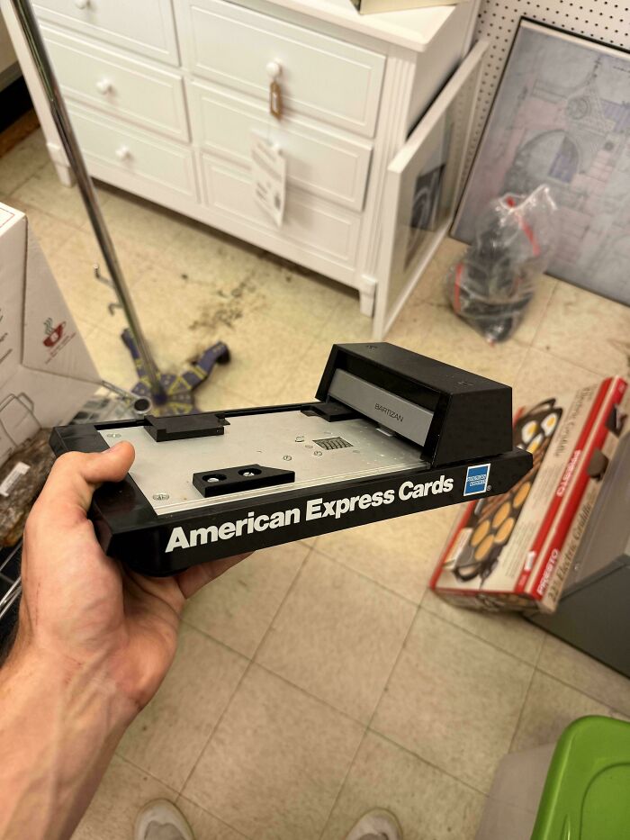 Hand holding a vintage American Express Cards reader device on a tiled floor with household items in background.